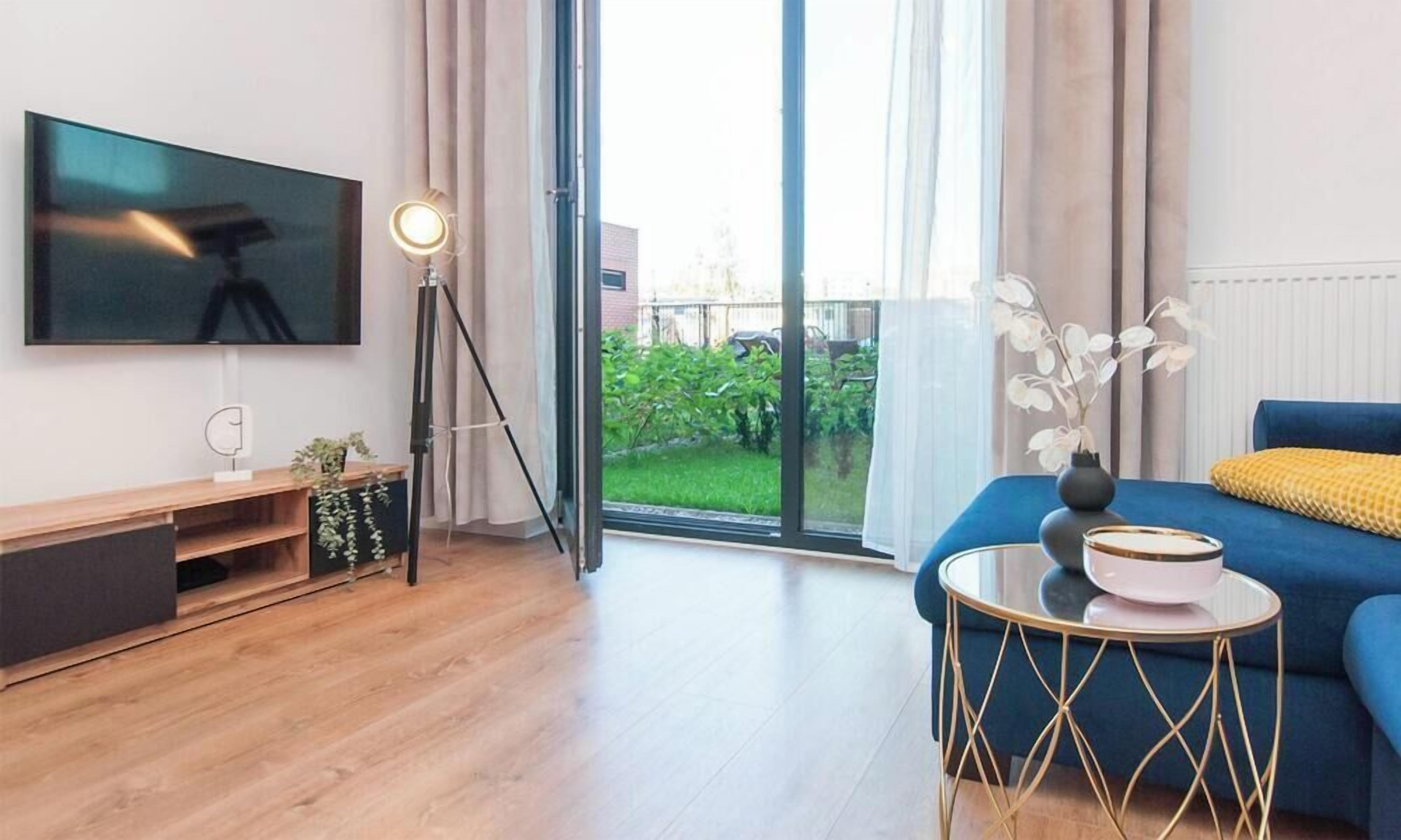Ein modernes Wohnzimmer mit einem Flachbildfernseher, einem Stativ, einem kleinen Tisch mit einer Schale, einem blauen Sofa und Blick auf einen grünen Garten durch eine Schiebetür.