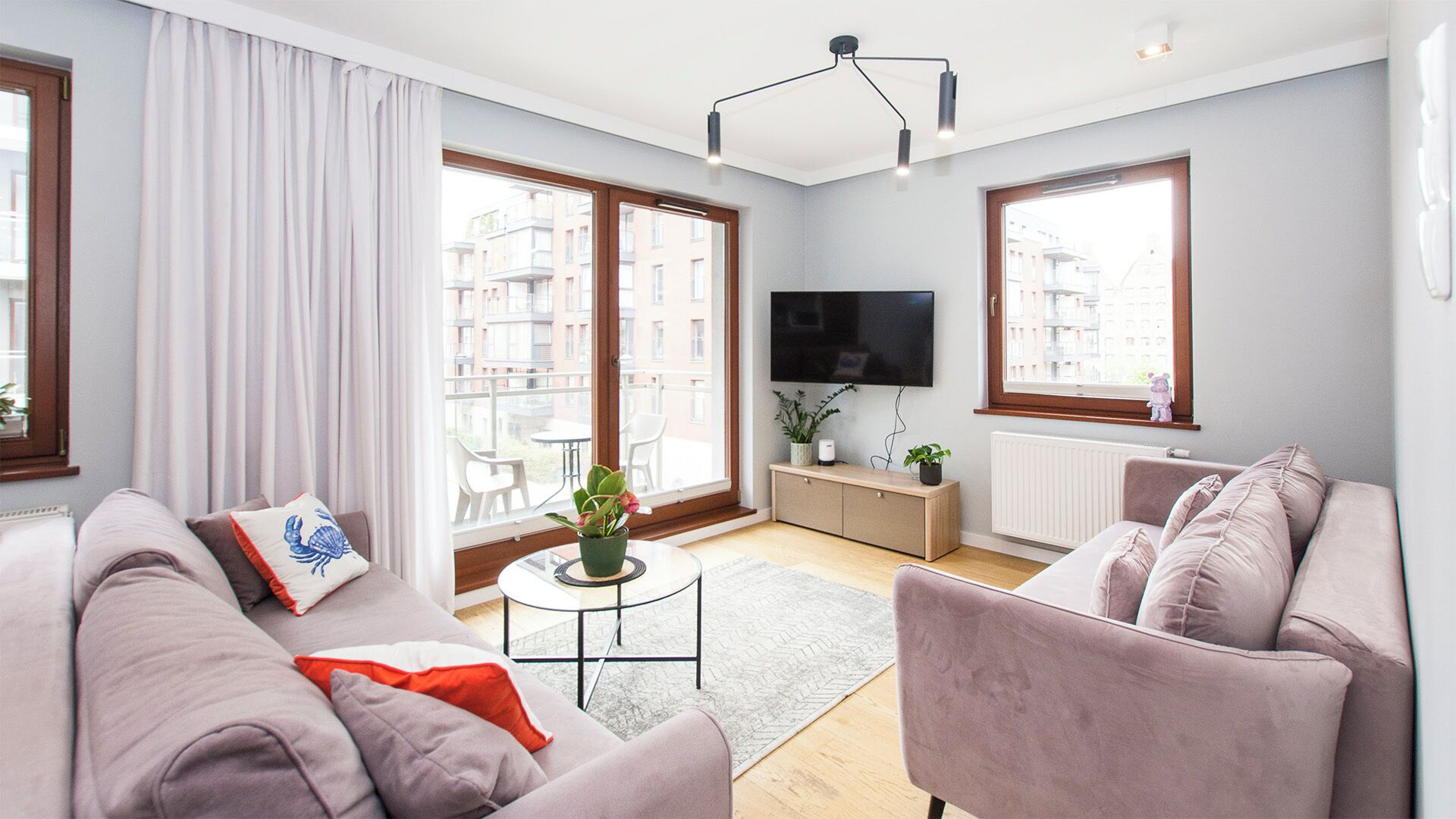 A modern living room with a sofa, a coffee table, and a TV. There are large windows with a view of buildings outside.
