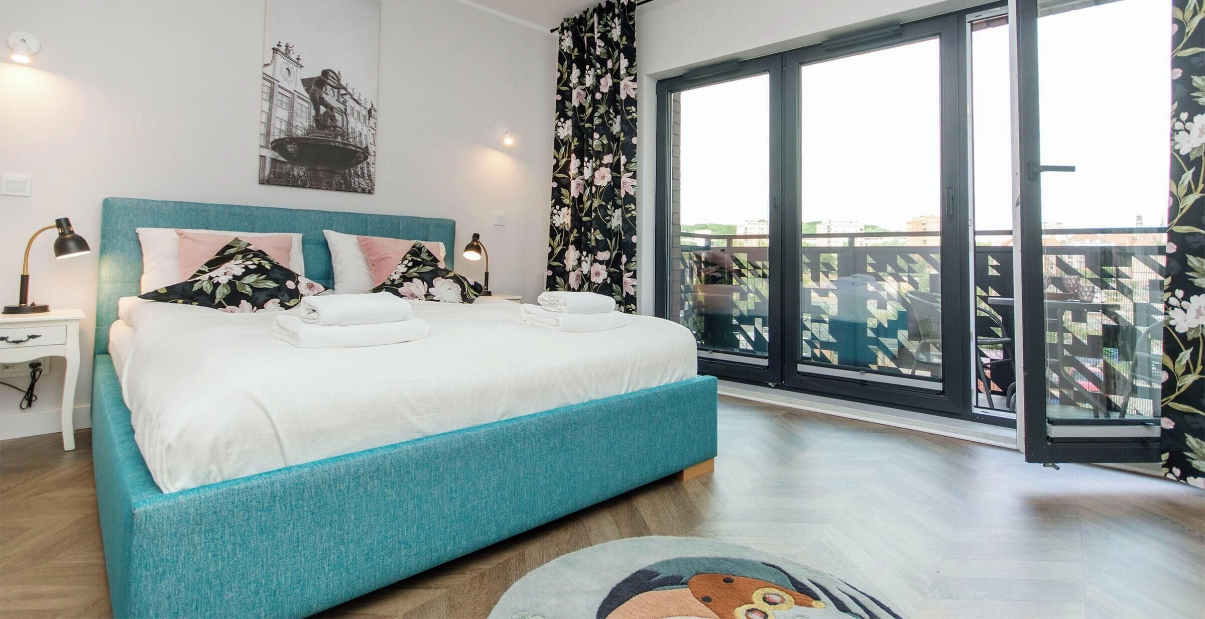 A bedroom with a bed, a nightstand, a lamp, a window with a view of buildings, and a balcony with a railing.