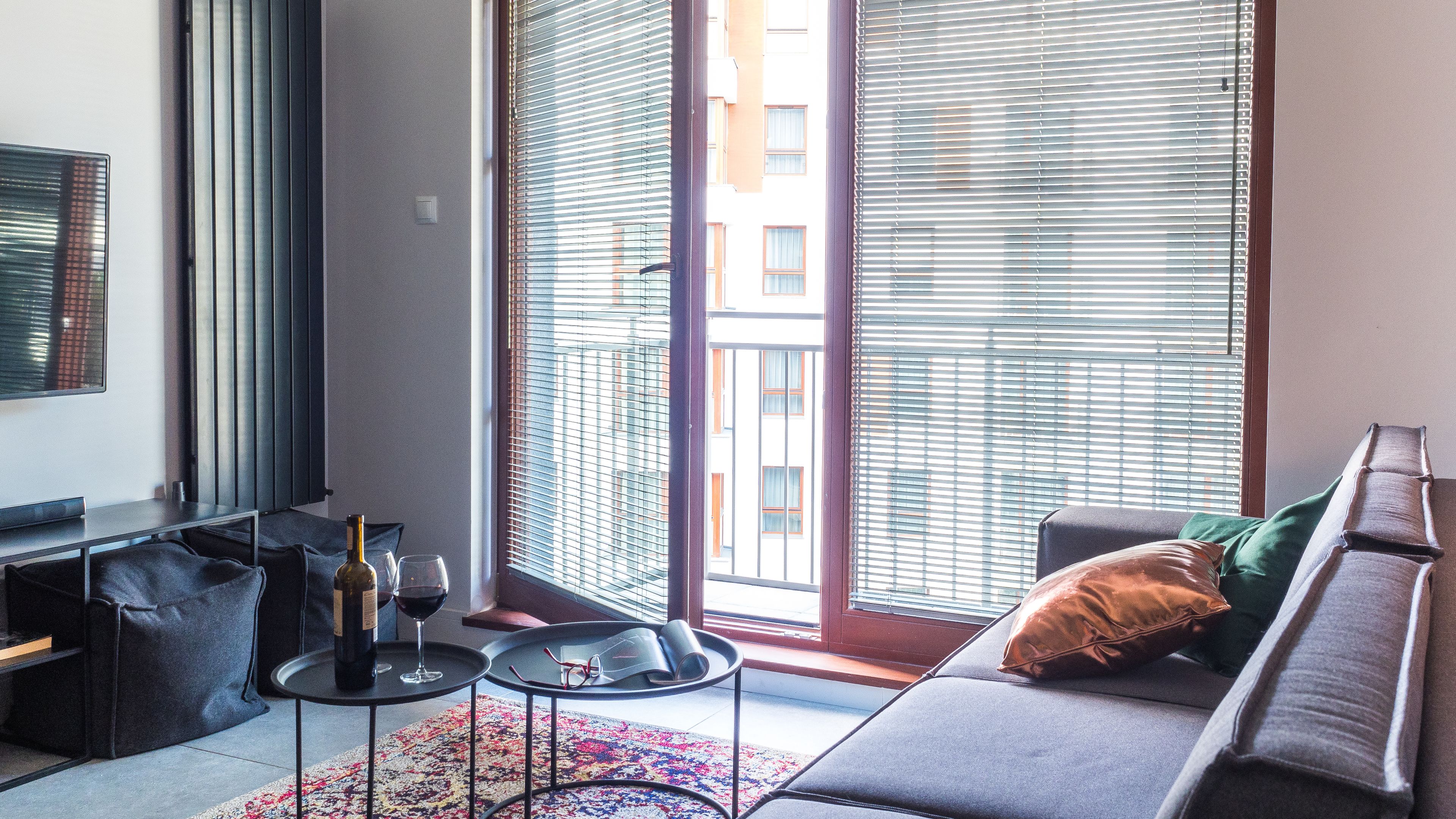 Een moderne woonkamer met een bank, een bijzettafeltje met een fles en glazen, en een groot raam met zonwering.