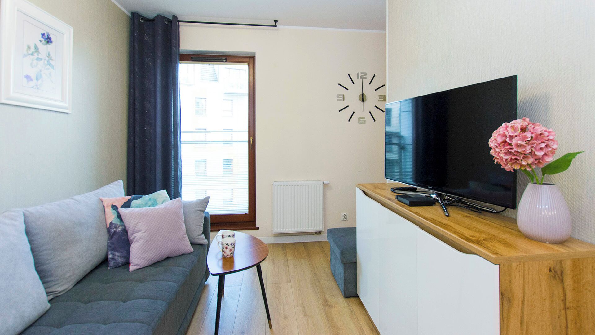 A modern living room with a sofa, a coffee table, a TV on a wooden stand, a clock, and a window with curtains.