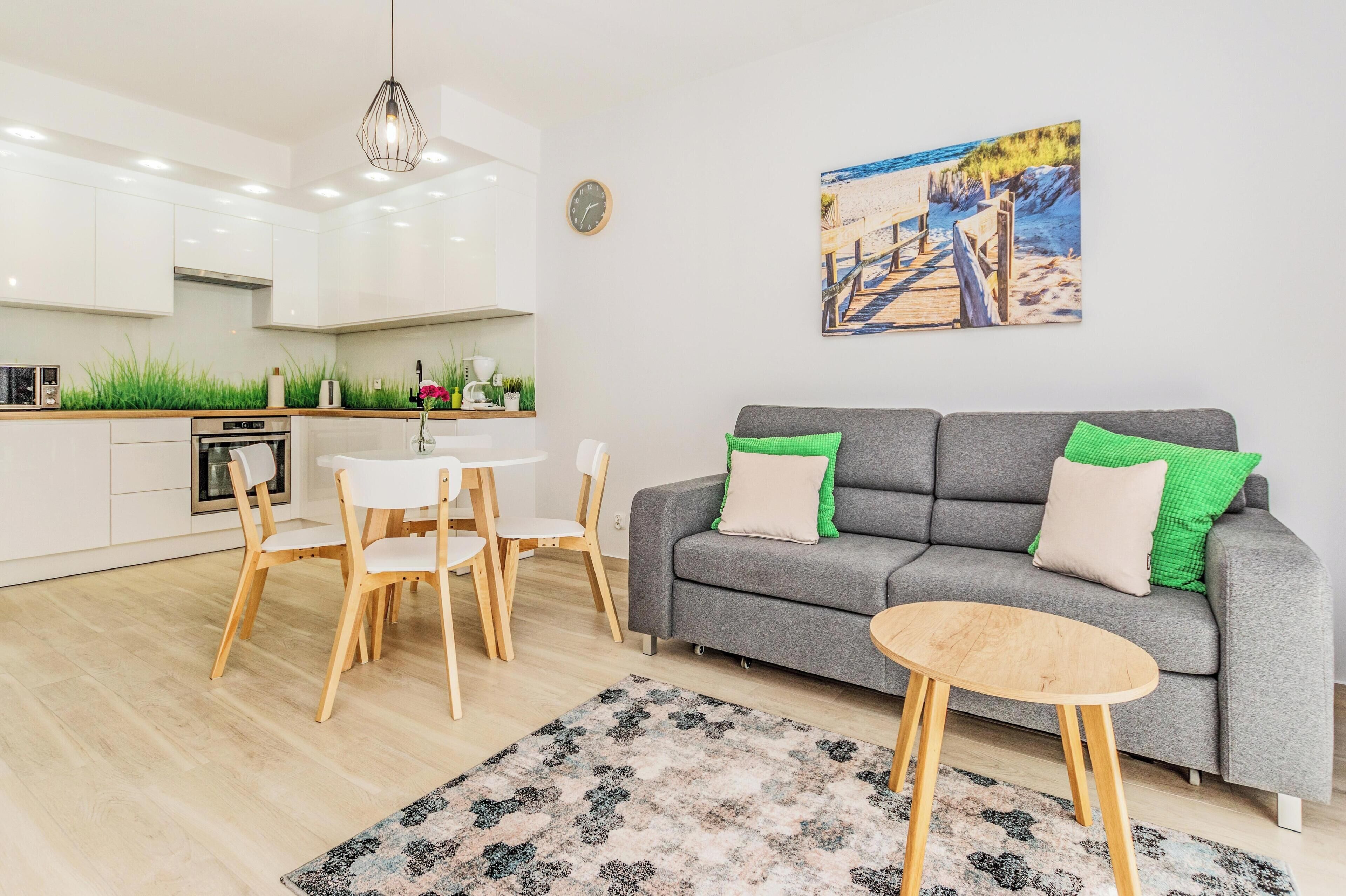 Une cuisine moderne avec des meubles blancs, un coin repas avec une table et des chaises, un canapé gris agrémenté de coussins verts, et un tableau sur le mur dont le thème est la plage.