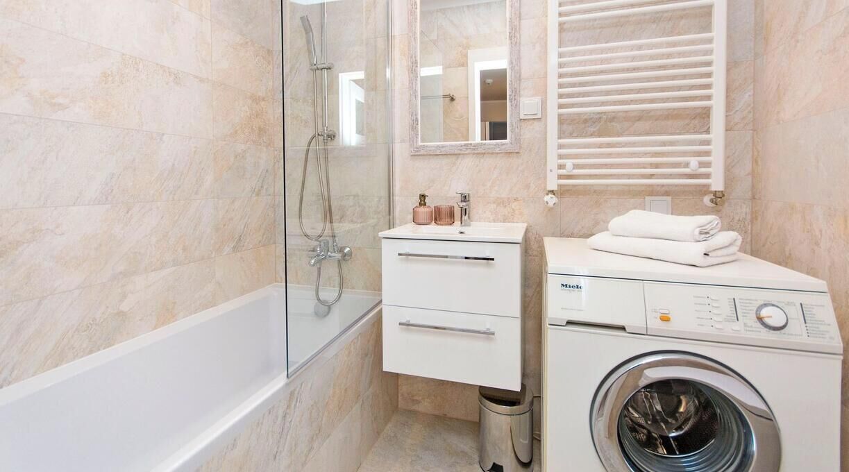 Ein Badezimmer mit einer gläsernen Dusche, einem weißen Waschbecken mit Spiegel, einem Handtuchhalter und einer Waschmaschine mit Frontlader.