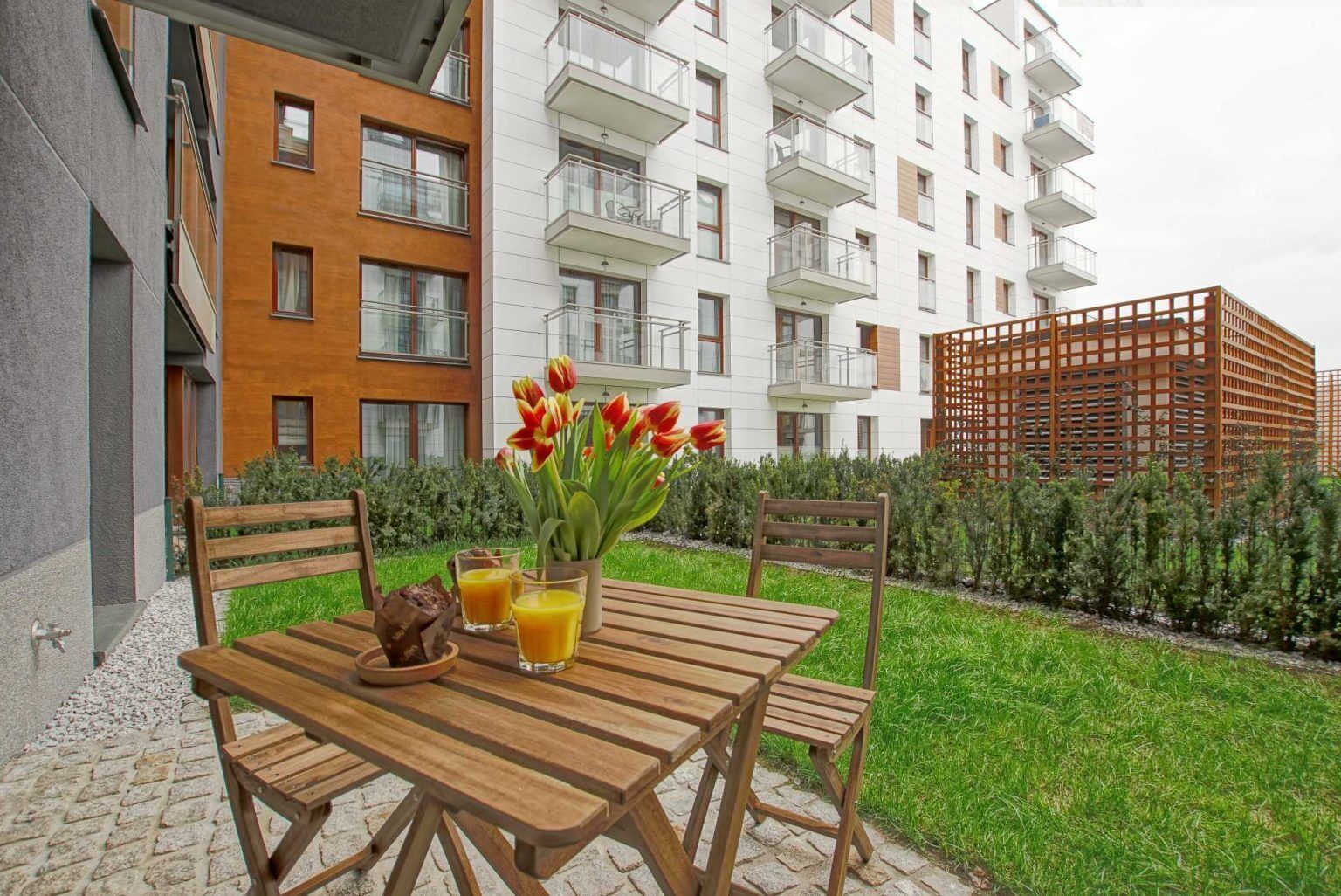 A wooden table with chairs, a vase of red tulips, and glasses of orange juice in an outdoor setting with a view of a modern apartment building.