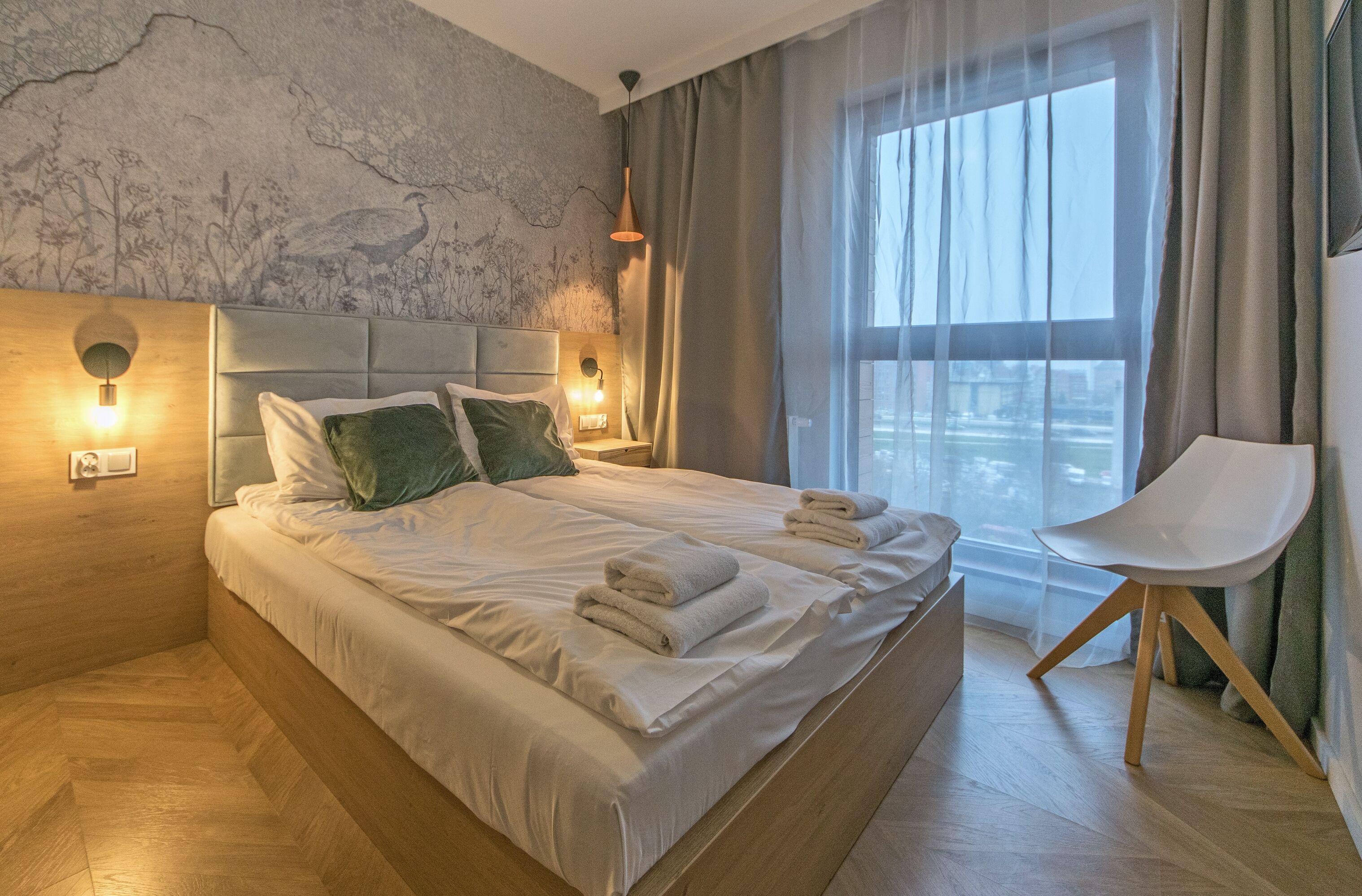 A modern hotel room with a large bed, two wall-mounted lights, a view of the city through sheer curtains, and a wooden floor.