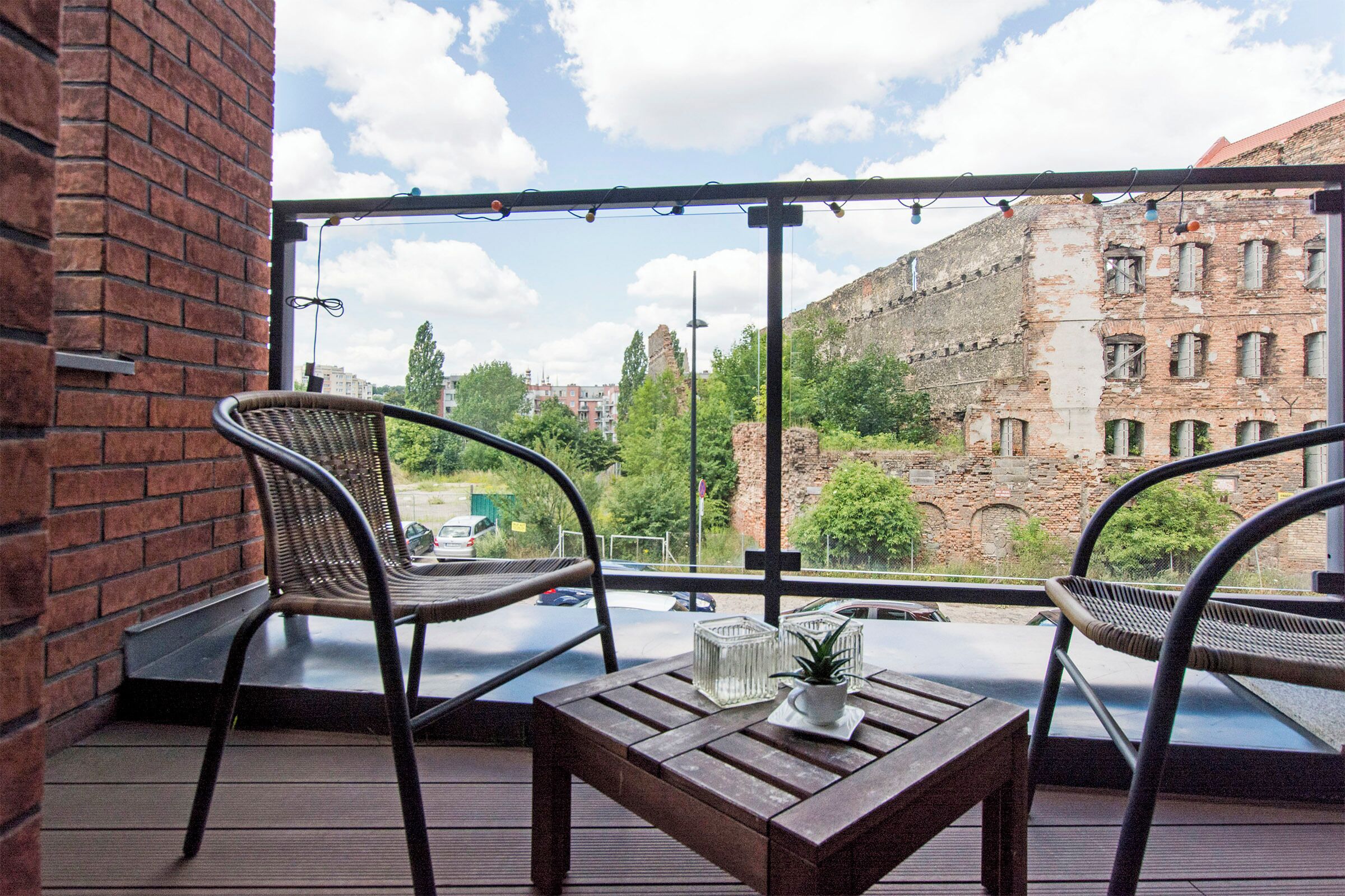 Un balcón con dos sillas de mimbre, una mesa de madera con una planta y vista a un edificio histórico de ladrillo.