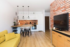 A modern kitchen with white cabinets, a brick accent wall, a flat-screen TV, and a yellow sofa.