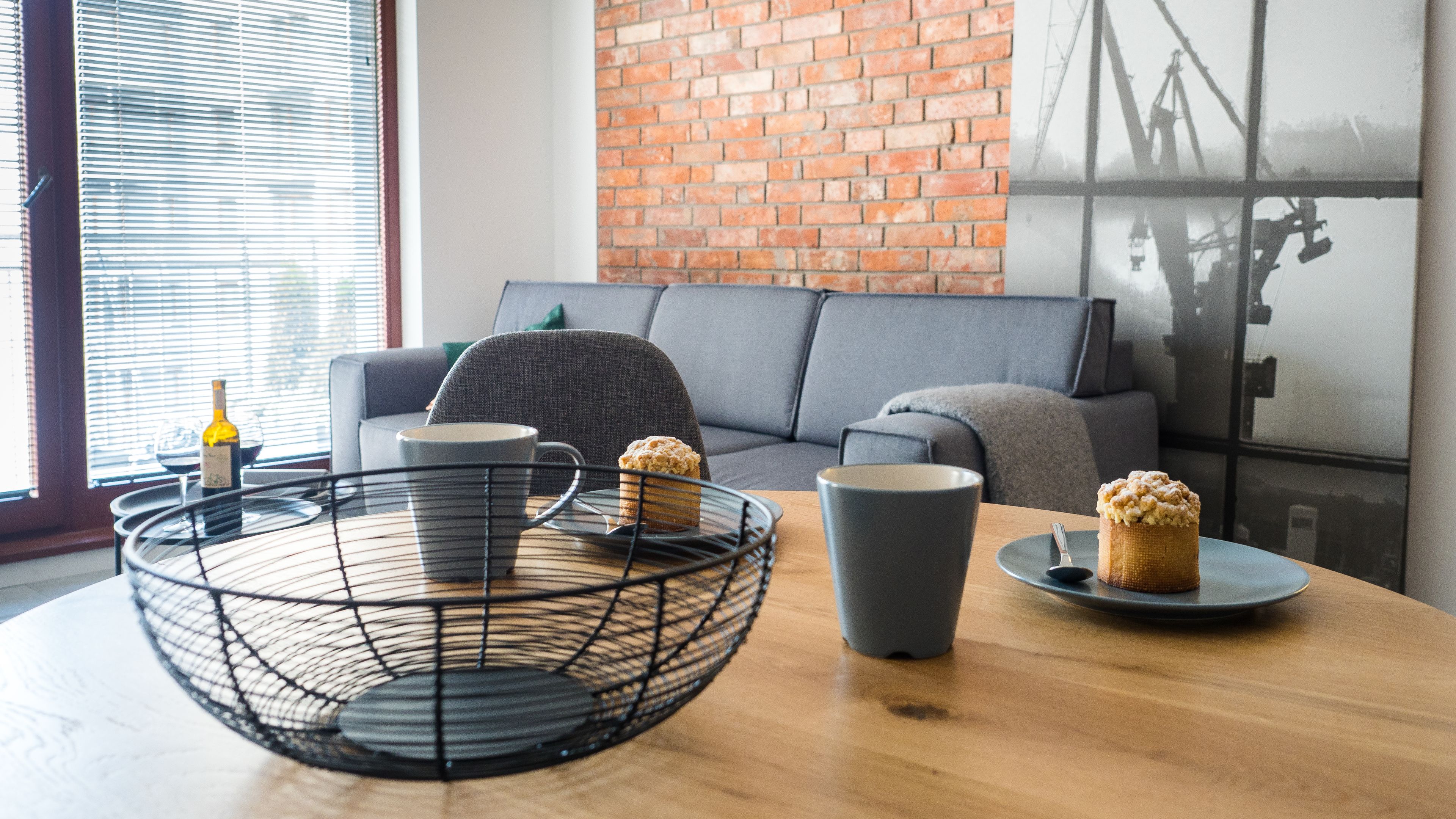 Un salon moderne avec un mur en briques, un canapé gris, une table basse avec un panier en fil de fer et une fenêtre avec des stores.