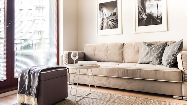 A living room with a beige sofa, two framed pictures on the wall, a small round table with a glass of wine, and a dark ottoman.