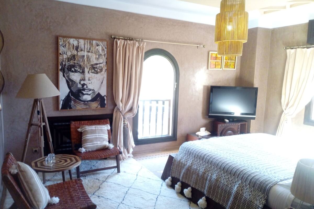 A bedroom with a bed, a television, a painting, a lamp, and a view of the outside.
