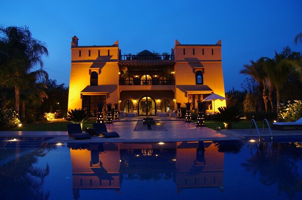 A large, two-story building with a pool in front, surrounded by palm trees and well-lit by ambient lighting. - Villa Malika Silvana (Marrakech)