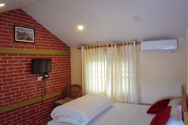 Una habitación de hotel con pared de ladrillo, un televisor montado en la pared, una cama con ropa blanca y almohadas rojas, una ventana con cortinas y un equipo de aire acondicionado.