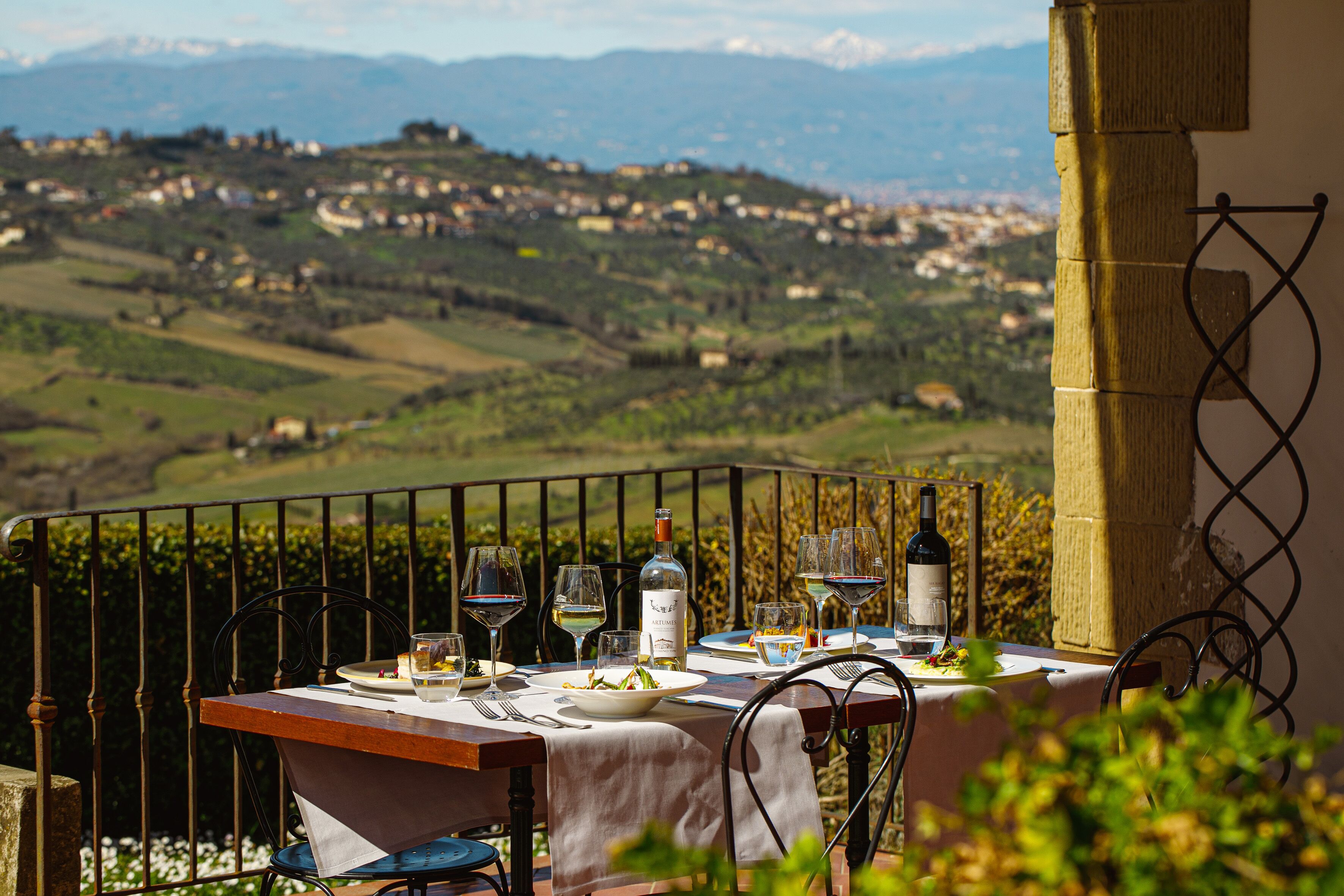 A table set for two with wine glasses, plates, and a bottle, overlooking a scenic landscape with hills and a village.