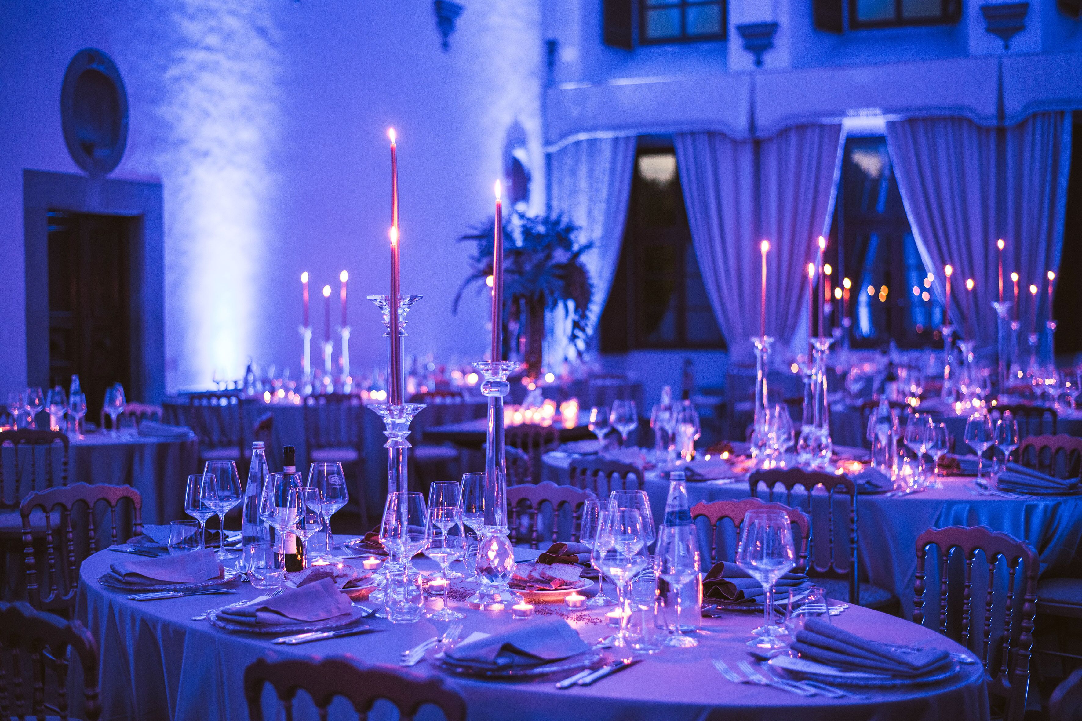 A banquet hall with round tables set for a formal event, featuring candlelit centerpieces and elegant table settings.