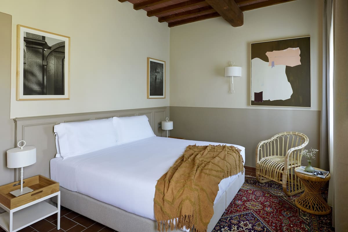 Une chambre d’hôtel bien aménagée, avec un grand lit, une table de chevet avec une lampe, une chaise et une petite table sur laquelle est posé un vase.