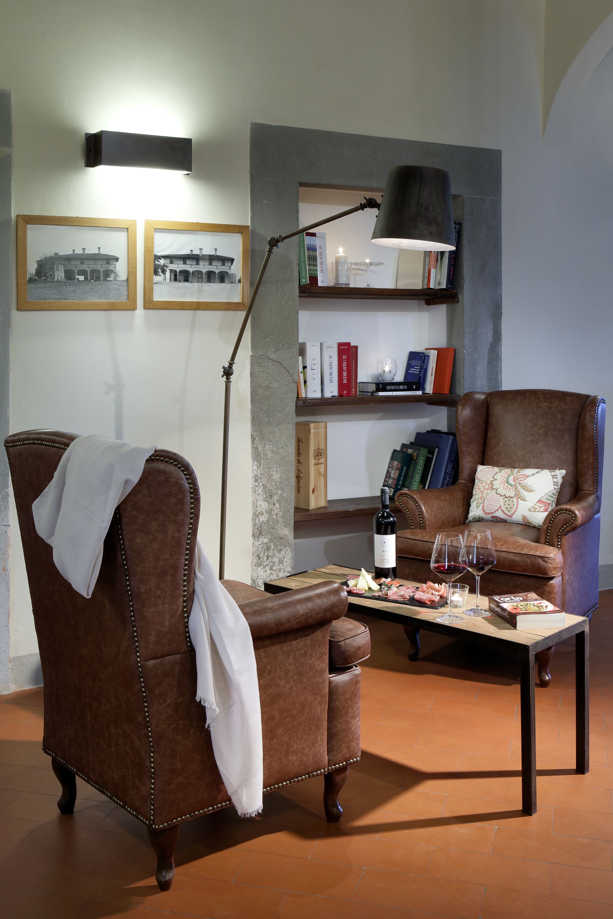 Una sala de estar acogedora con un sillón de cuero marrón, un sofá a juego, una mesa de centro con aperitivos y una botella, una lámpara de pie y una estantería con libros y adornos.