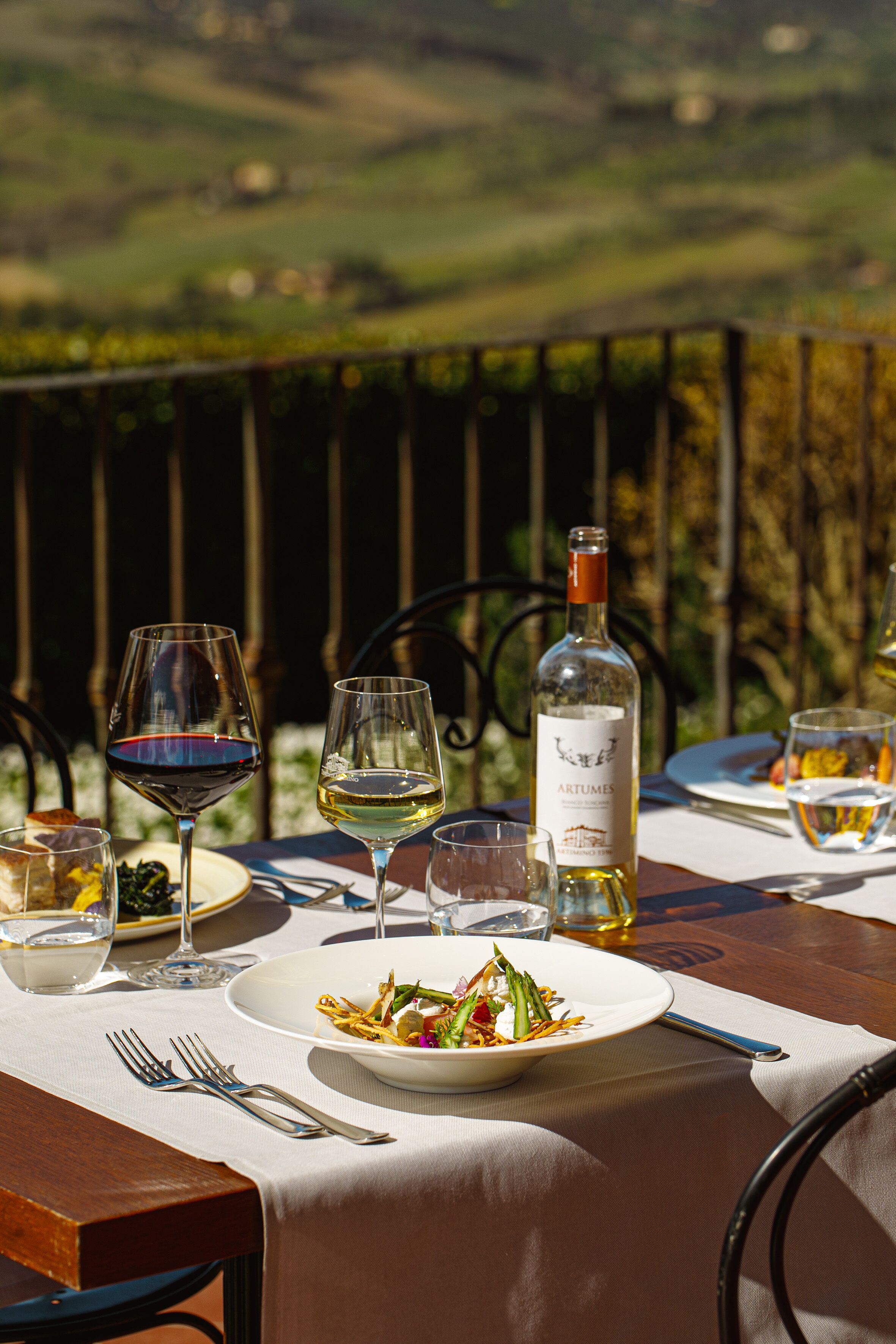 Una mesa de comedor dispuesta con platos, vasos y una botella de vino, con vistas a un paisaje pintoresco.