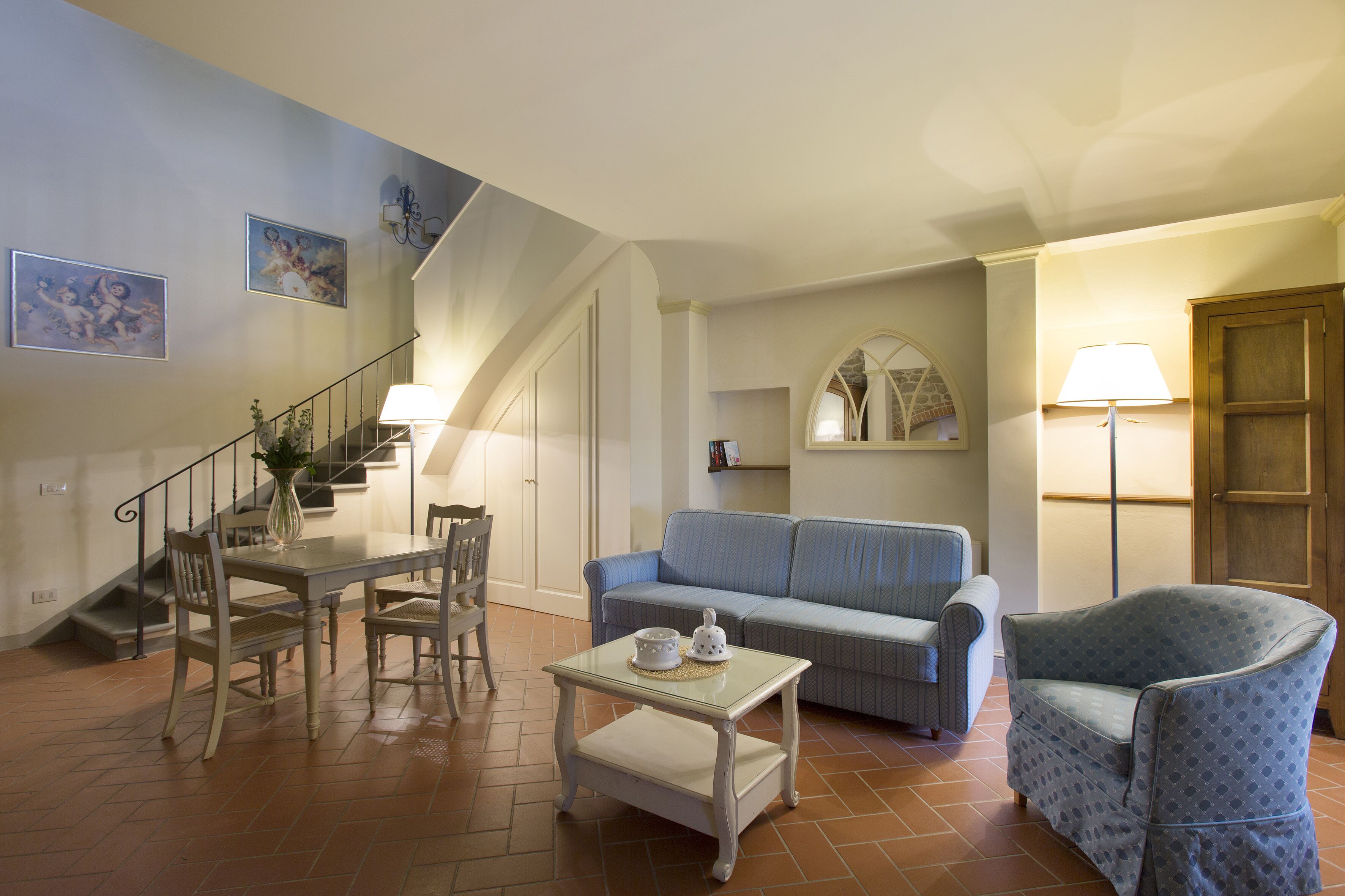 A living room with a dining table, chairs, a sofa, a patterned armchair, a staircase, and a lamp.