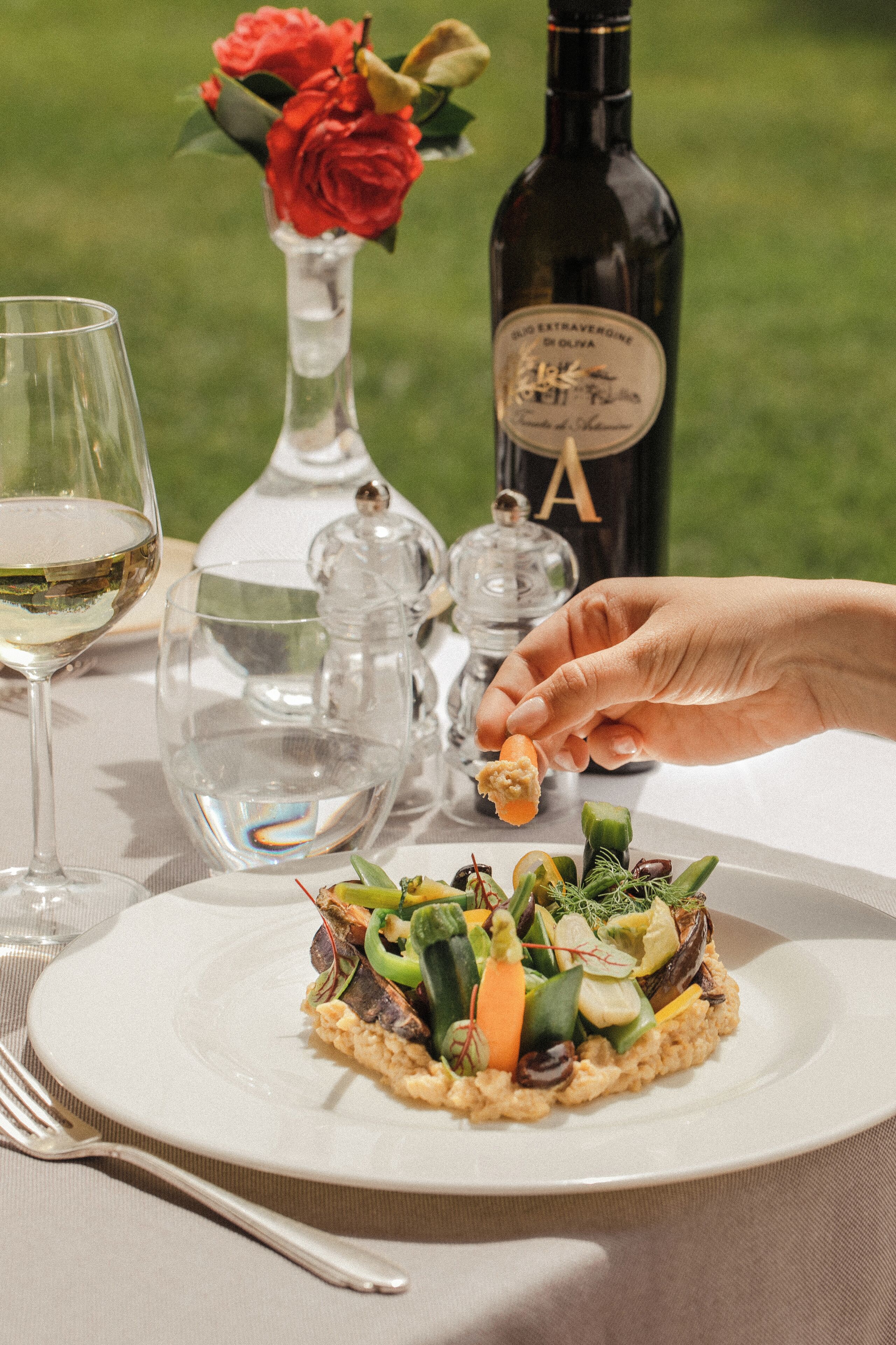 A dining table with a plate of food, a bottle of wine, a glass of wine, a vase with flowers, and a salt and pepper shaker.