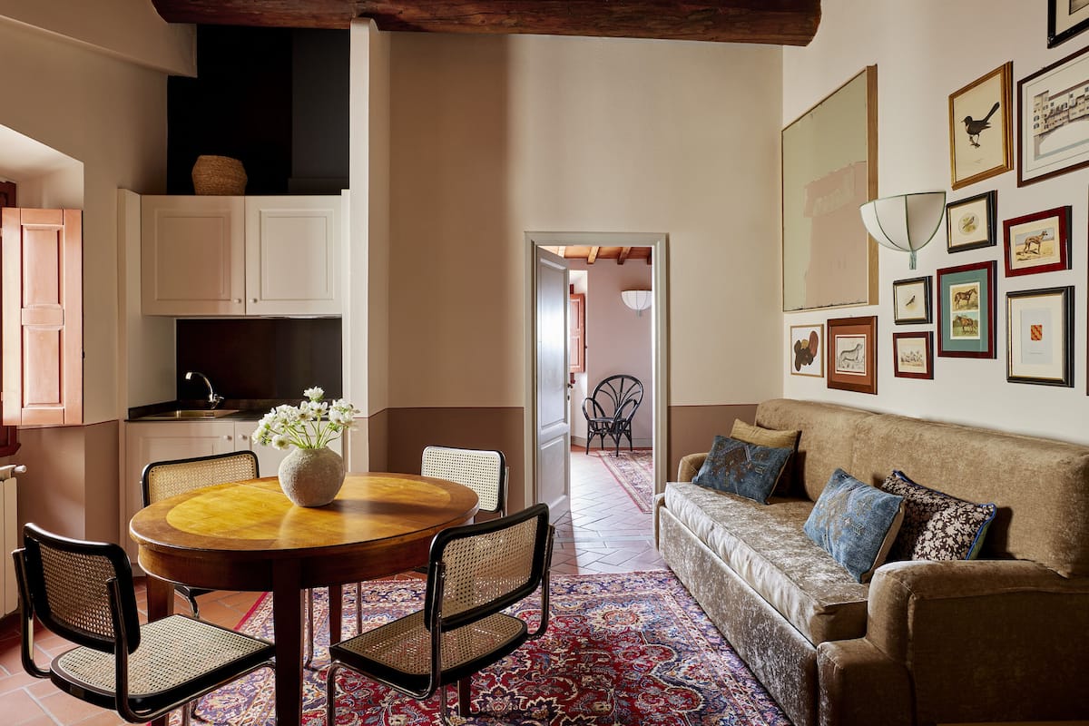 Un coin repas avec une table ronde, des chaises et un vase de fleurs. On y trouve également un canapé avec des coussins et un mur orné de tableaux.