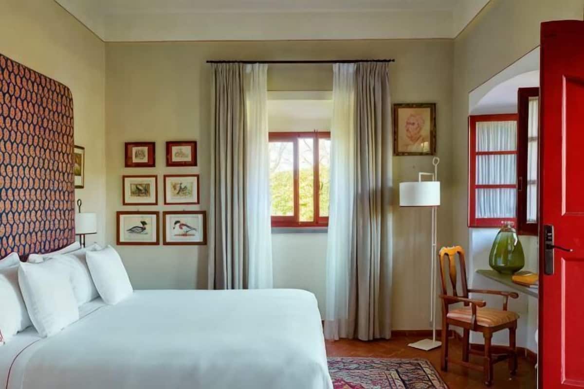 Une chambre à coucher avec un lit, une chaise, une lampe, une fenêtre avec des rideaux et des tableaux encadrés accrochés au mur.