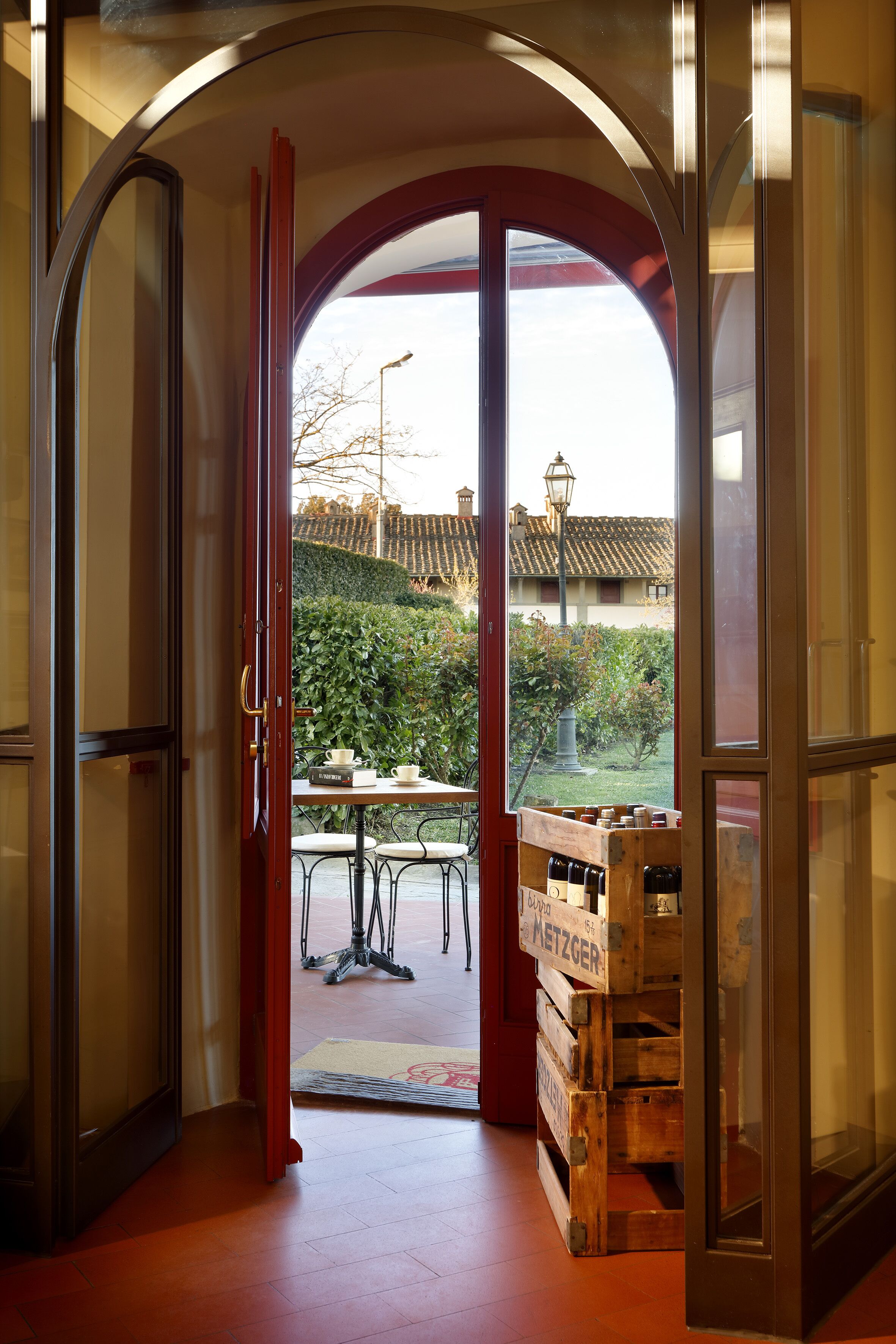 Eine geöffnete Tür führt zu einer Terrassenfläche mit Tisch und Stühlen, einer Holzkiste mit Flaschen sowie einem Blick auf einen Garten und Gebäude im Hintergrund.