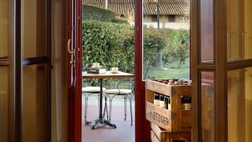 Eine geöffnete Tür führt zu einer Terrassenfläche mit Tisch und Stühlen, einer Holzkiste mit Flaschen sowie einem Blick auf einen Garten und Gebäude im Hintergrund.