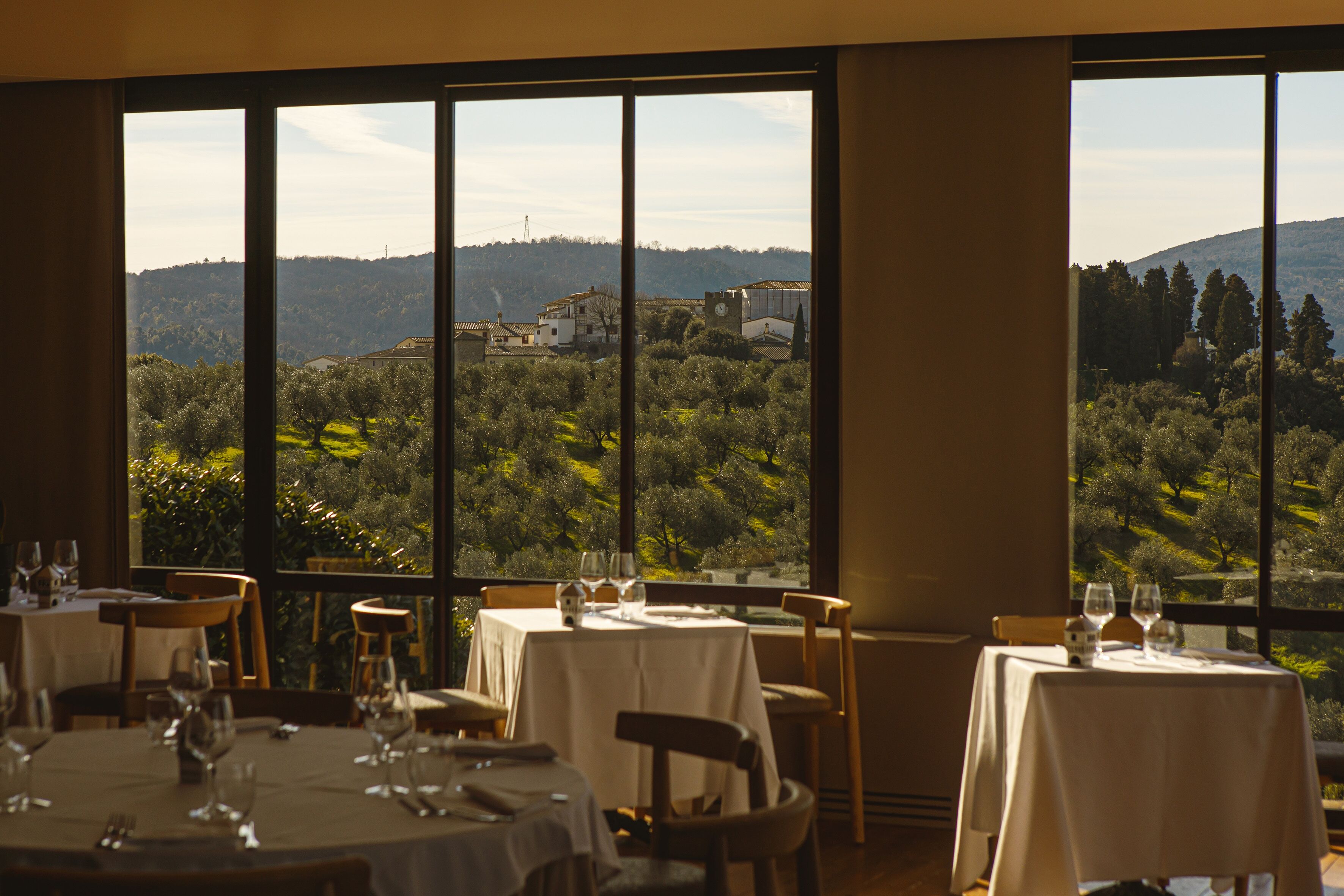 Un espace repas avec des tables préparées pour deux personnes, de grandes fenêtres offrant une vue sur un paysage vallonné.