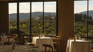 A dining area with tables set for two, large windows offering a view of a hilly landscape.