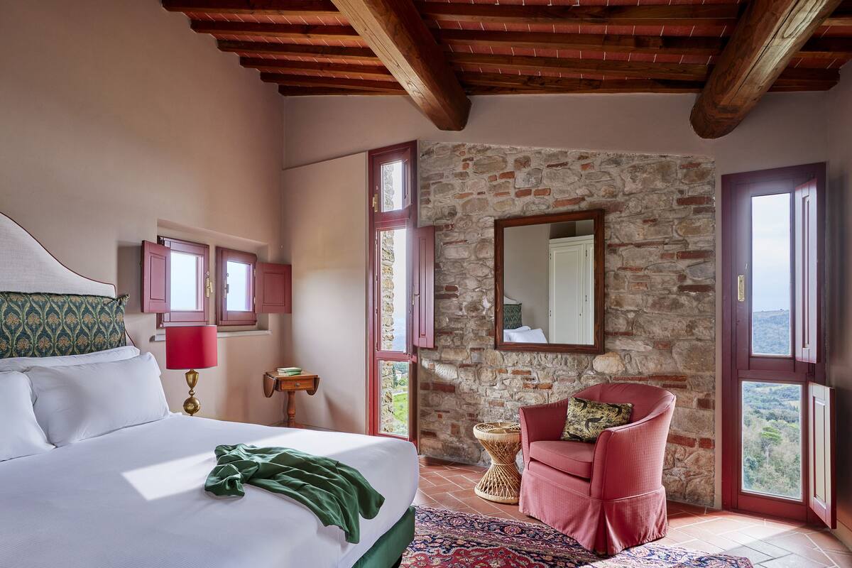 Une chambre avec un grand lit, un mur en pierre, un fauteuil rouge et une vue par la fenêtre.
