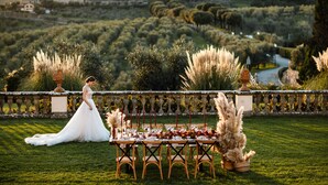 Une décoration de mariage comprenant une mariée, des tables ornées de centres de table floraux et un décor pittoresque composé d’un vignoble et de bâtiments.