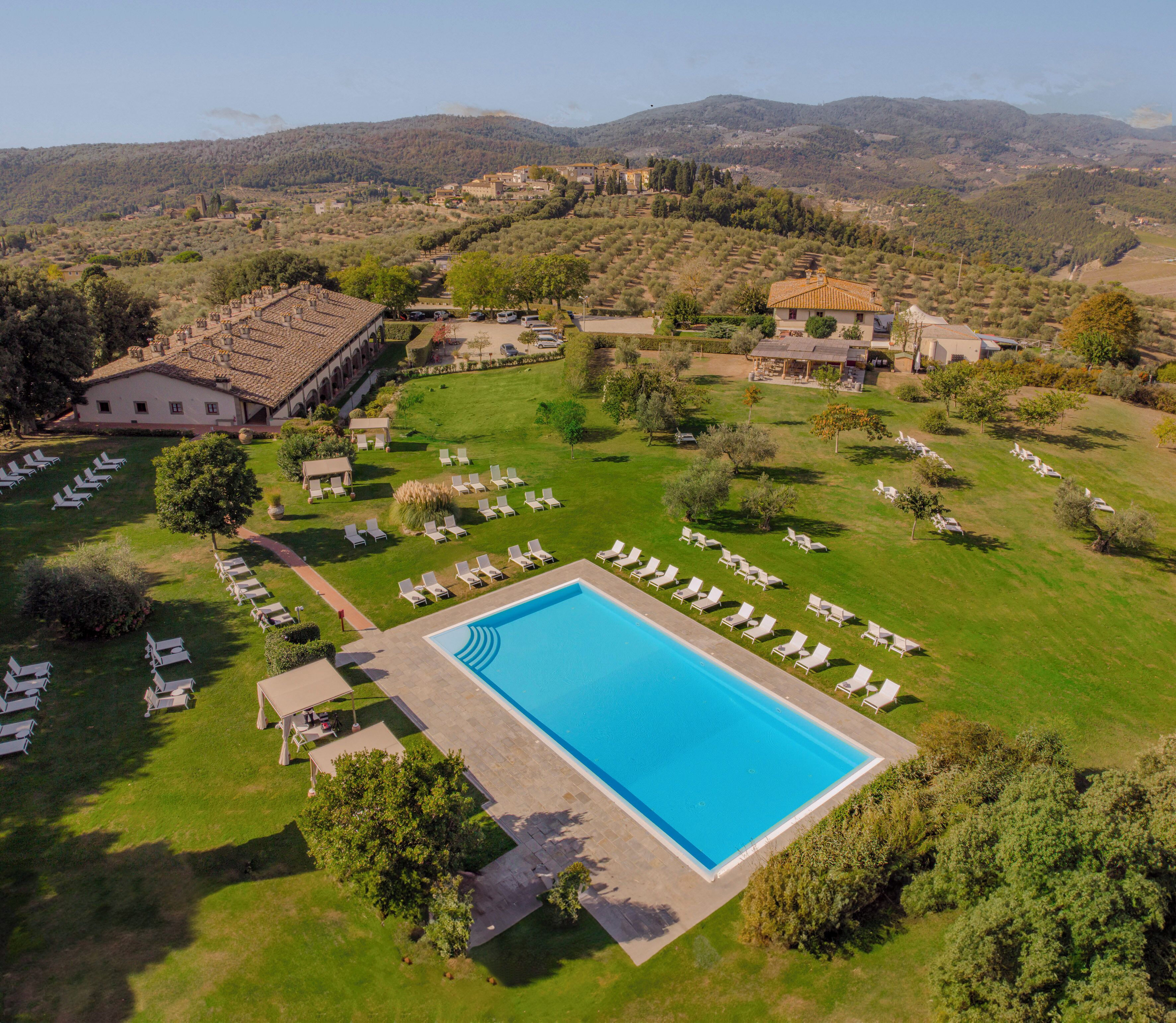 An aerial view of a resort with a swimming pool surrounded by lounge chairs, a large building, and a mountainous backdrop.