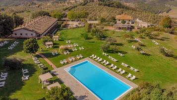 An aerial view of a resort with a swimming pool surrounded by lounge chairs, a large building, and a mountainous backdrop.