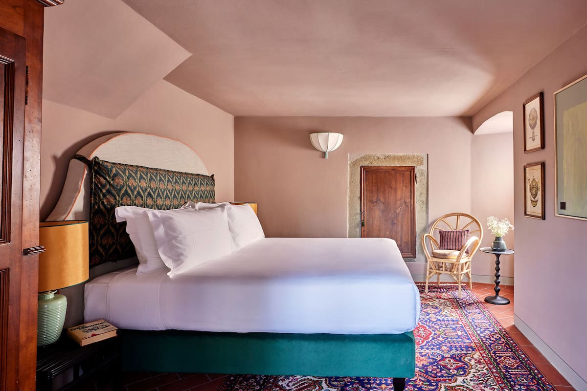 Un lit bien fait, avec du linge de lit blanc, une tête de lit à motifs et un couvre-lit vert. Une petite table avec une lampe et un livre se trouve à côté du lit. Une chaise est placée près d’une porte ornée d’un cadre décoratif.