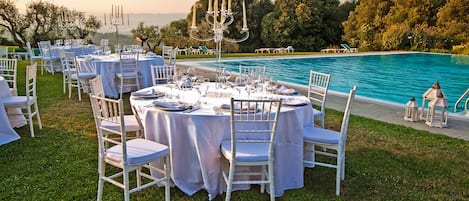 An outdoor dining area with round tables and white chairs, set against a backdrop of a swimming pool and lush greenery.