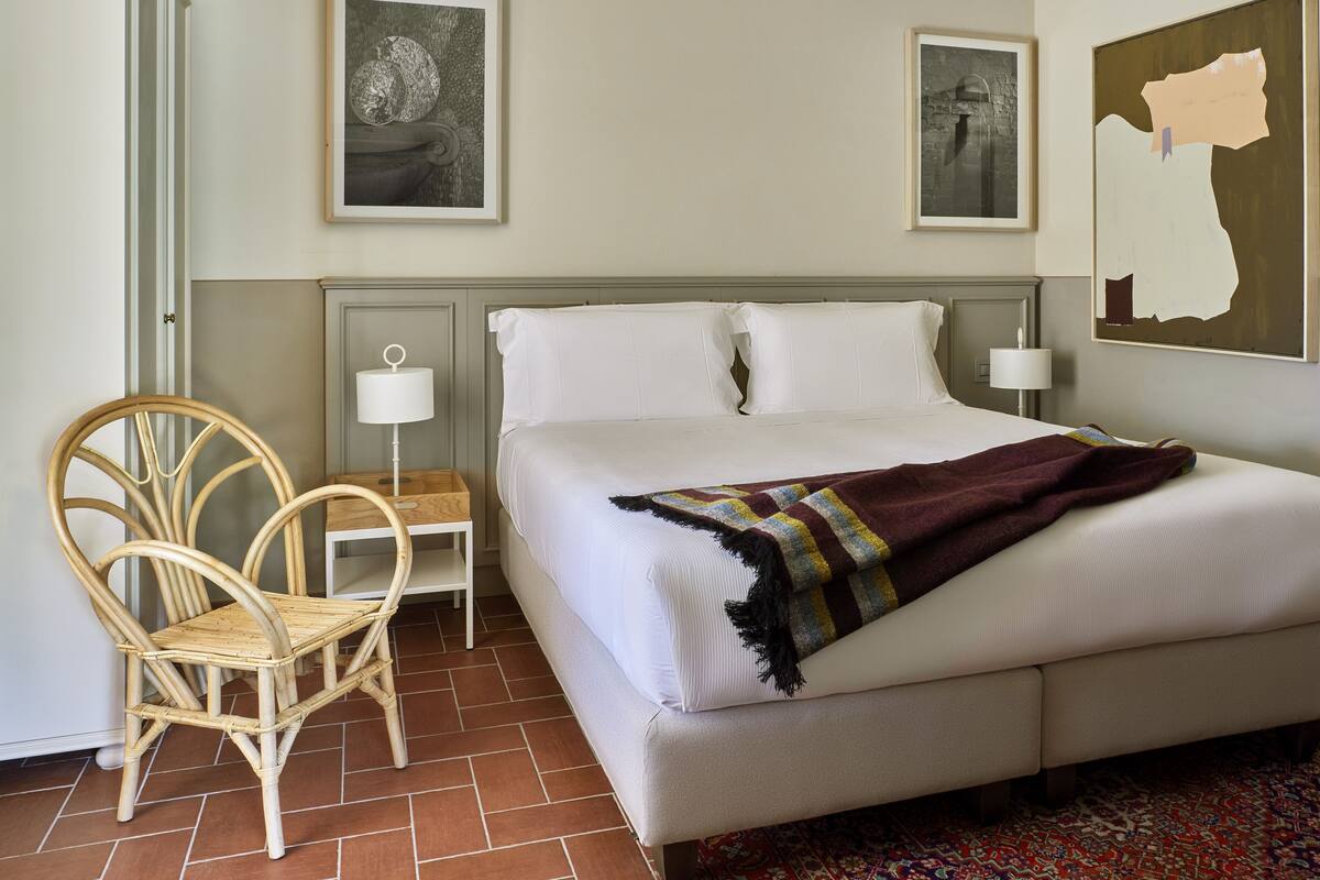 Une chambre d’hôtel bien rangée, avec un lit, une chaise, une table de chevet et deux lampes fixées au mur.