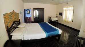 A hotel room with a bed, a desk, a chair, a small table, a mirror, a window with curtains, and a ceiling with recessed lighting. - Pavithram Homestay (Devikolam)