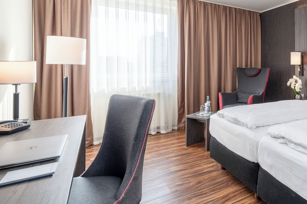 Ein Hotelzimmer mit Schreibtisch, zwei Betten, einem Sessel, einem Nachttisch und einer Lampe.