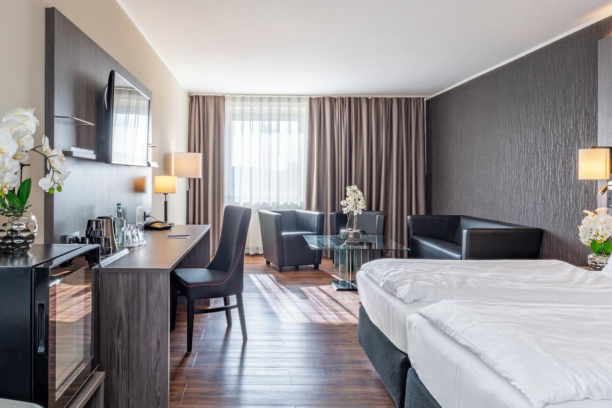 Ein Hotelzimmer mit Bett, Schreibtisch mit Stuhl, Sofa, Fernseher und einem Fenster mit Vorhängen.