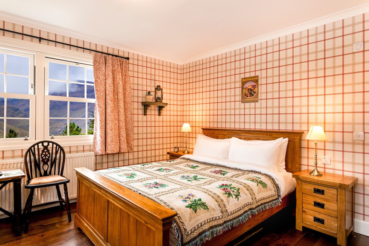 Un dormitorio con cama de madera, mesita de noche con lámpara, una silla, ventana con cortinas y pared con estampado de cuadros.