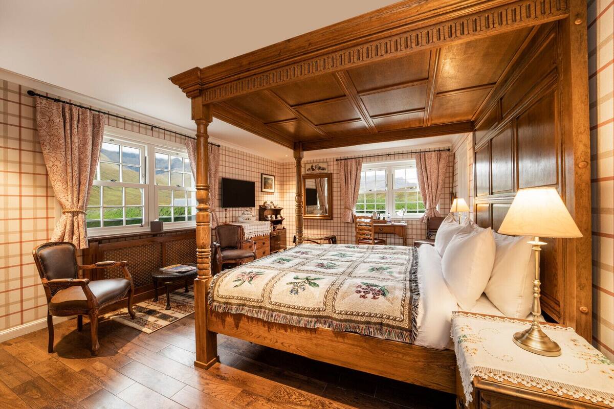 Una cama con dosel tradicional, con cubrecama floral, un escritorio de madera con una silla, y vistas al exterior a través de dos ventanas.