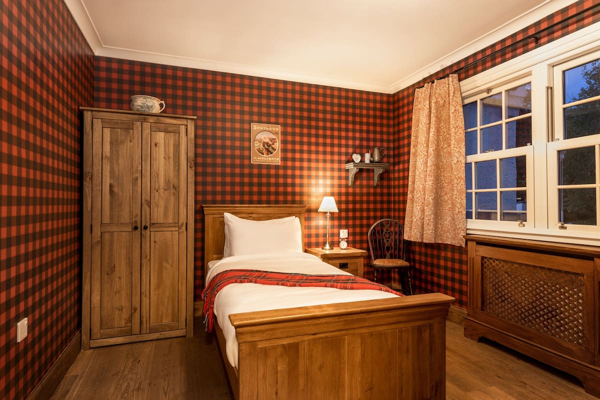 Una habitación con papel tapiz a cuadros, una cama de madera, una mesita de noche con una lámpara, una silla y una ventana con cortinas.