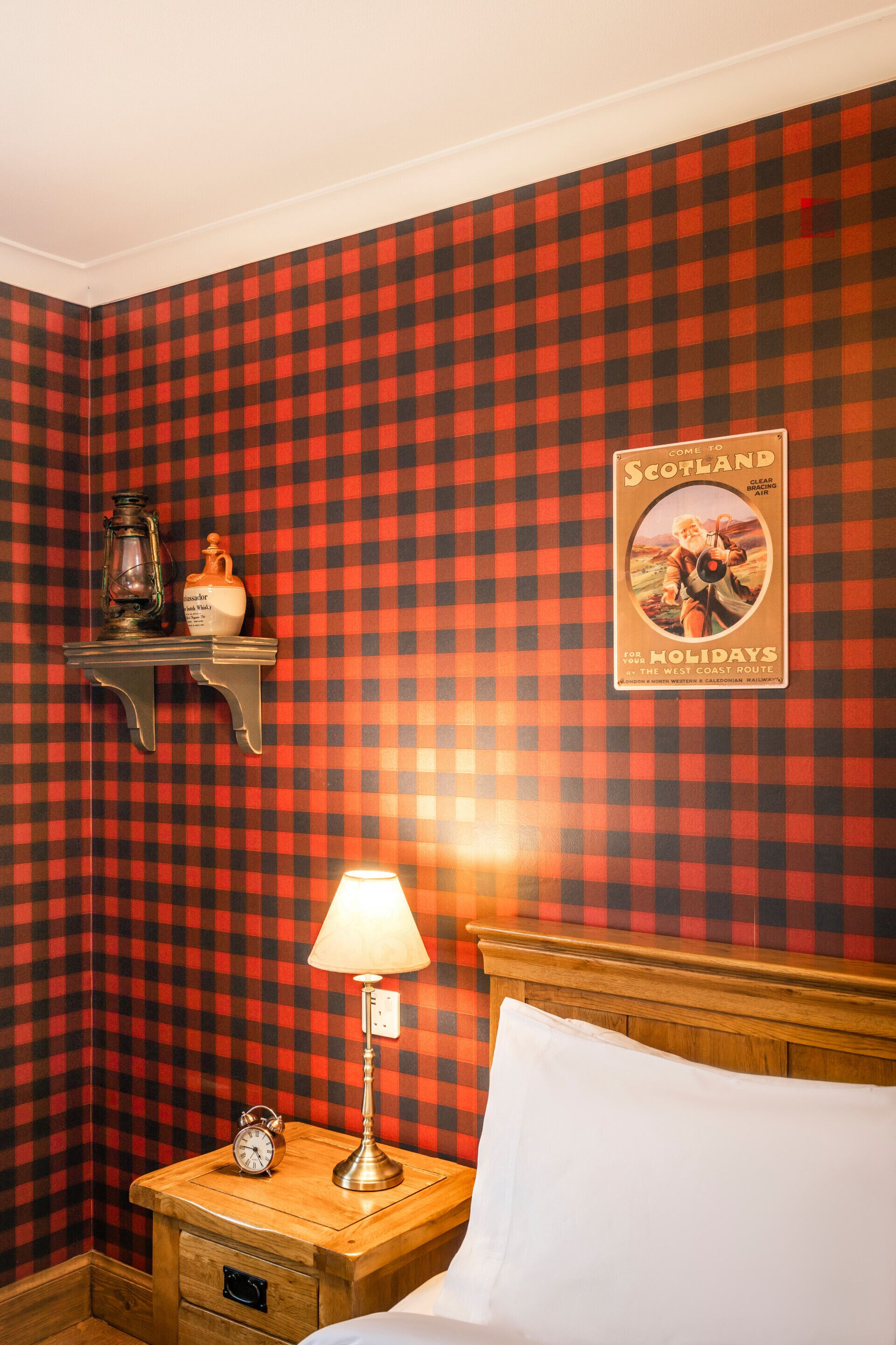 A room with a checkered wall, a nightstand with a lamp, a clock, and a shelf with a bottle and a lantern.