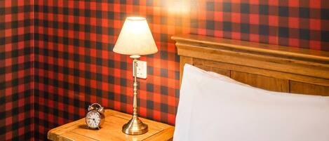 A room with a checkered wall, a nightstand with a lamp, a clock, and a shelf with a bottle and a lantern.