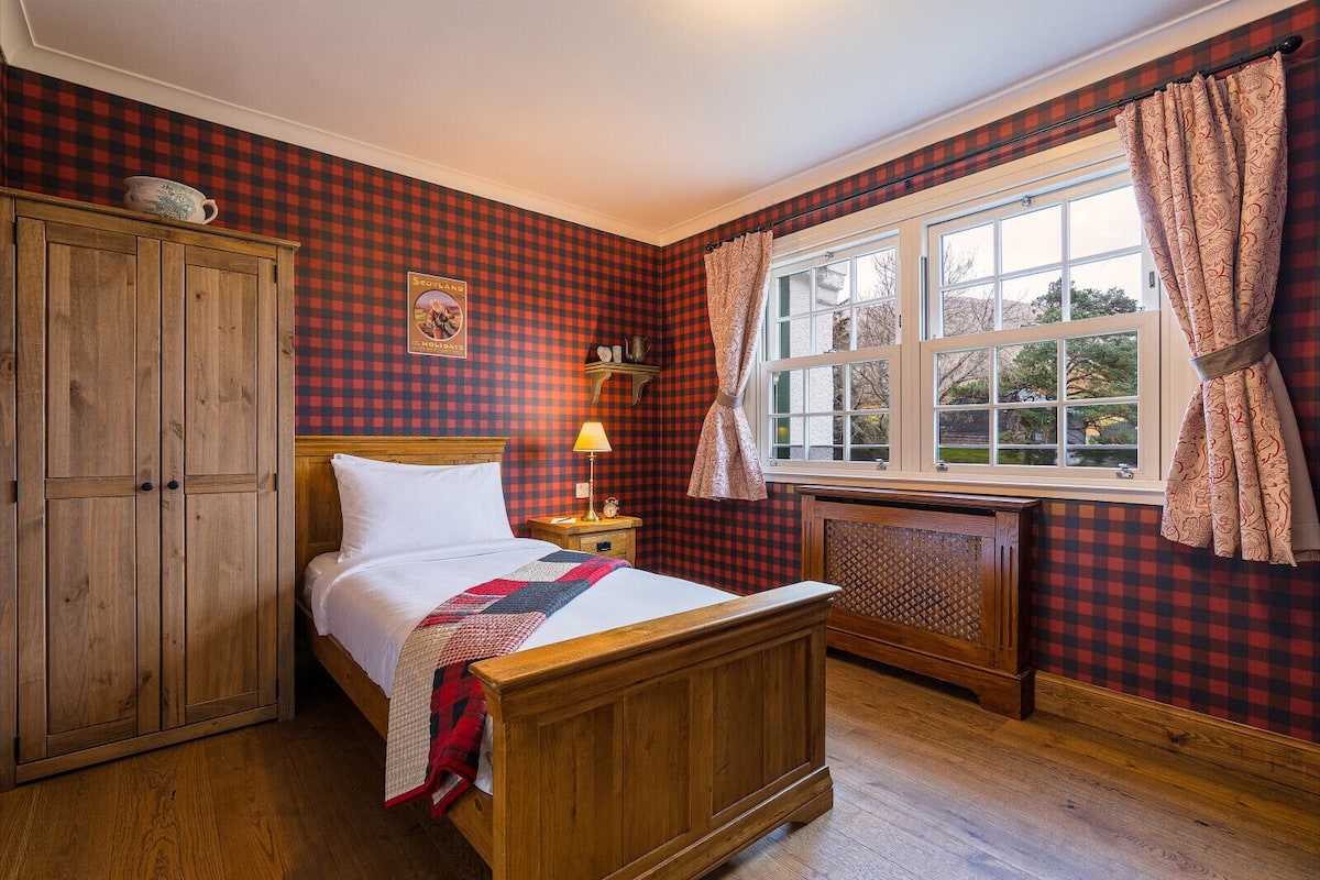 Una habitación tradicional con una cama de madera, una mesita de noche con una lámpara, una ventana con cortinas y un papel tapiz a cuadros.