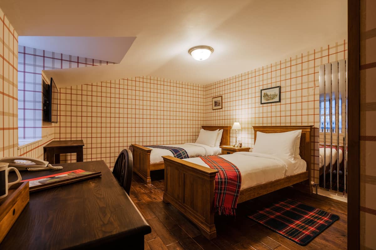 Una habitación de hotel con dos camas de madera, un escritorio y un teléfono, y una alfombra de cuadros.