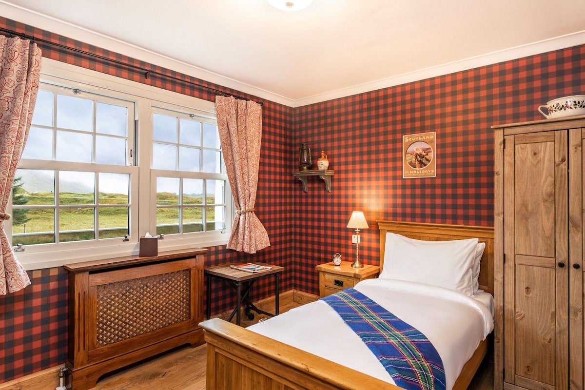 A room with a bed, a wooden wardrobe, a nightstand with a lamp, a window with curtains, and a view of the outdoors.