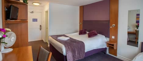 Een hotelkamer met een bed, een televisie op een plank, een ventilator en een spiegel.
