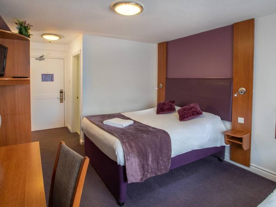 Hotellihuone, jossa on sänky, hyllyllä oleva televisio, tuuletin ja peili.