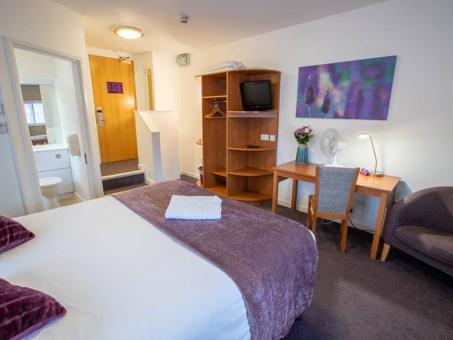 A hotel room with a bed, a desk with a chair, a television, a fan, and a painting on the wall.